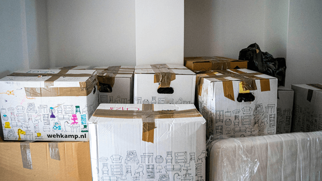 This image shows several cardboard boxes stacked in a room. The boxes are taped and feature drawings or illustrations of furniture and household items, possibly indicating their contents. One of the boxes has a visible logo from "wehkamp.nl," which could be a reference to a company or brand. Additionally, there is a mattress or large padded item partially visible in the bottom of the image. The setting suggests that these boxes are part of a move or delivery process.