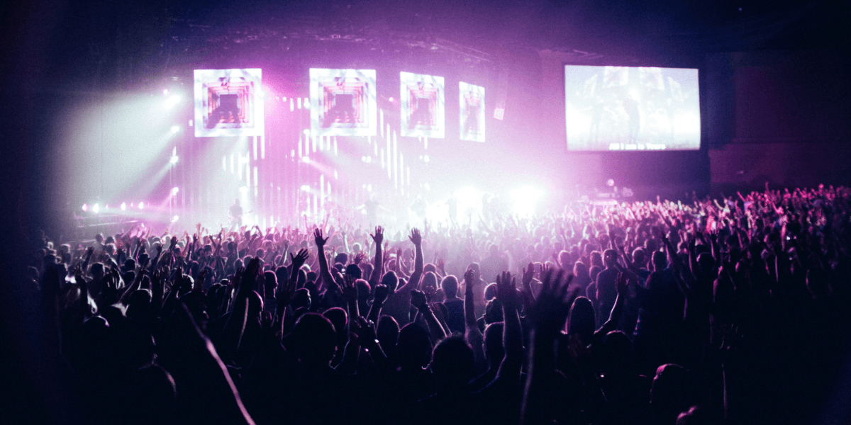 Crowd with raised hands at a vibrant concert, colorful lights illuminating the stage