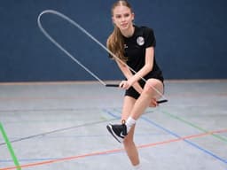 A person in black sports attire skillfully jump ropes indoors on a court. The blue wall and colorful court lines accentuate the action and focus.