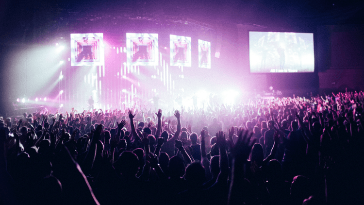 Crowd with raised hands at a vibrant concert, colorful lights illuminating the stage