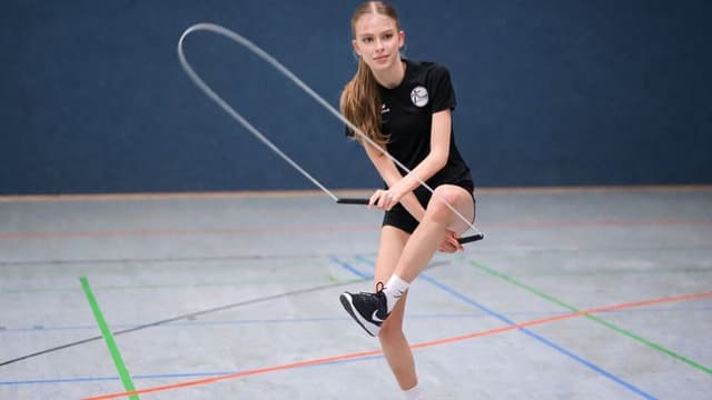 A person in black sports attire skillfully jump ropes indoors on a court. The blue wall and colorful court lines accentuate the action and focus.