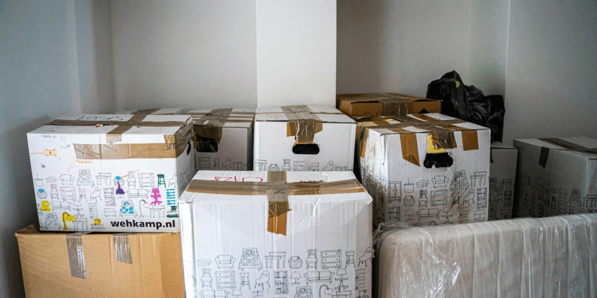 This image shows several cardboard boxes stacked in a room. The boxes are taped and feature drawings or illustrations of furniture and household items, possibly indicating their contents. One of the boxes has a visible logo from "wehkamp.nl," which could be a reference to a company or brand. Additionally, there is a mattress or large padded item partially visible in the bottom of the image. The setting suggests that these boxes are part of a move or delivery process.